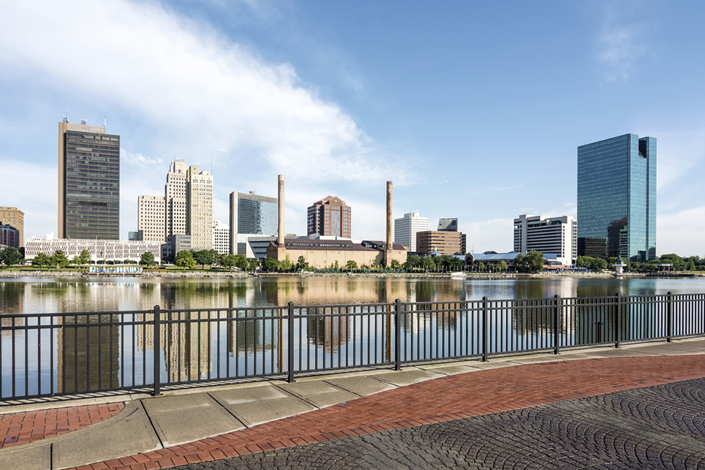 A  panoramic view of downtown Toledo Ohio's skyline from across the Maumee river at a popular restaurant area with a paver brick boardwalk and a decorative iron railing..  A beautiful  blue sky with white clouds for a backdrop.